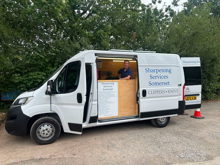 A white commercial van parked outdoors on a gravel surface with trees in the background. The side doors are open, revealing a wooden counter and a man standing inside.