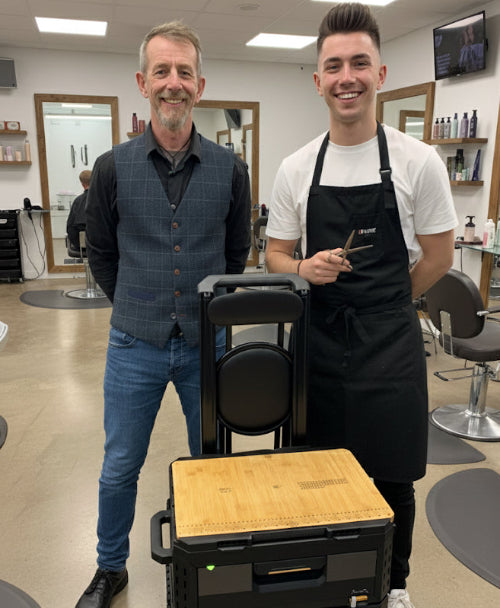 Two men standing in a salon with a chair and tools, one wearing an apron.