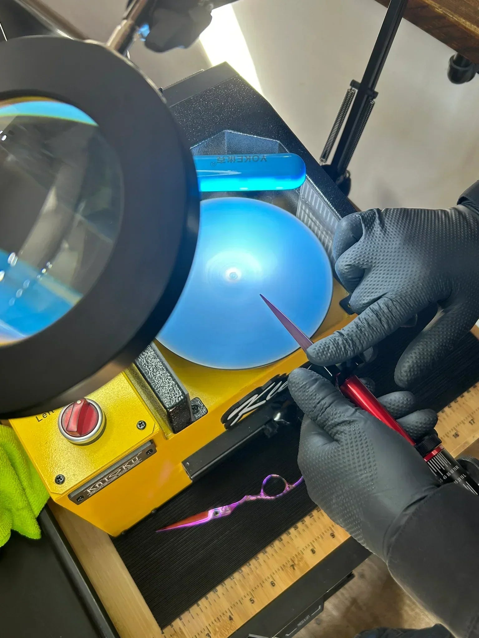 Close-up of a professional sharpening station, showing a gloved hand sharpening Hairdressor scissors on a blue rotating disc. A magnifying glass hovers above the disc for precision, with a ruler, purple scissors, and a blue "YORKER" cylinder nearby on the well-lit workbench.